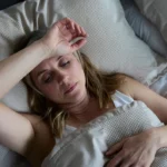 15 Early Signs of Perimenopause You Shouldn’t Ignore 1 A middle-aged woman lying in bed with her hand on her forehead, looking distressed.