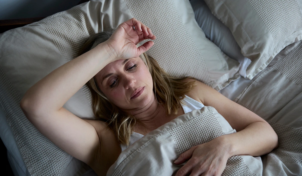 A middle-aged woman lying in bed with her hand on her forehead, looking distressed.